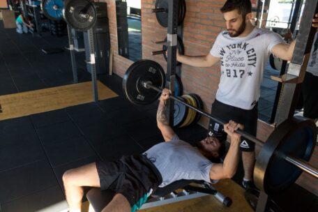 Personal trainer spotting a man performing a barbell bench press properly in a gym to ensure safe weightlifting technique.