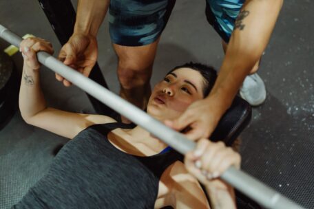 Personal trainer spotting a woman learning how to bench press properly with a barbell in a gym setting.
