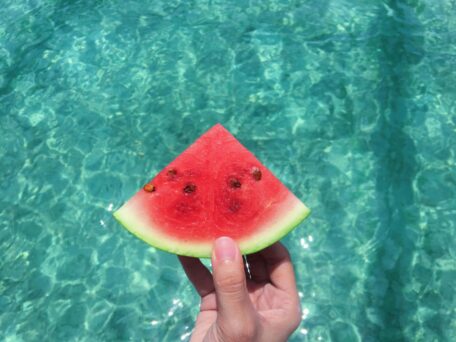 Stay hydrated: Hand holding a refreshing watermelon slice near crystal clear blue water. Summer hydration tip.