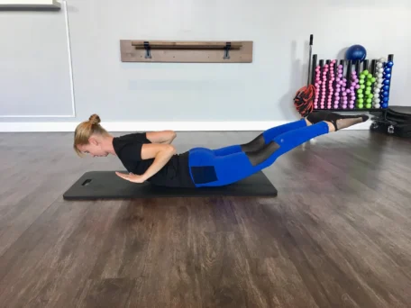 Woman doing prone back extension exercise to improve posture on a mat in a gym.