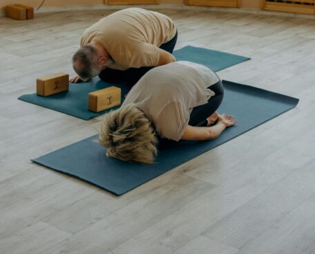 Two people doing child's pose exercise for posture improvement on yoga mats with blocks in a bright studio.