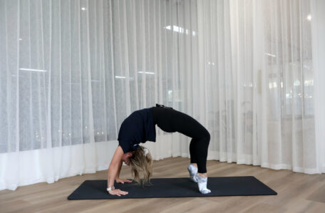 Woman performing a wheel pose yoga stretch on a black mat in front of sheer white curtains.