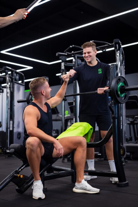 Personal trainer in a Club Lime shirt fist-bumps a man sitting on a weight bench in a modern gym.