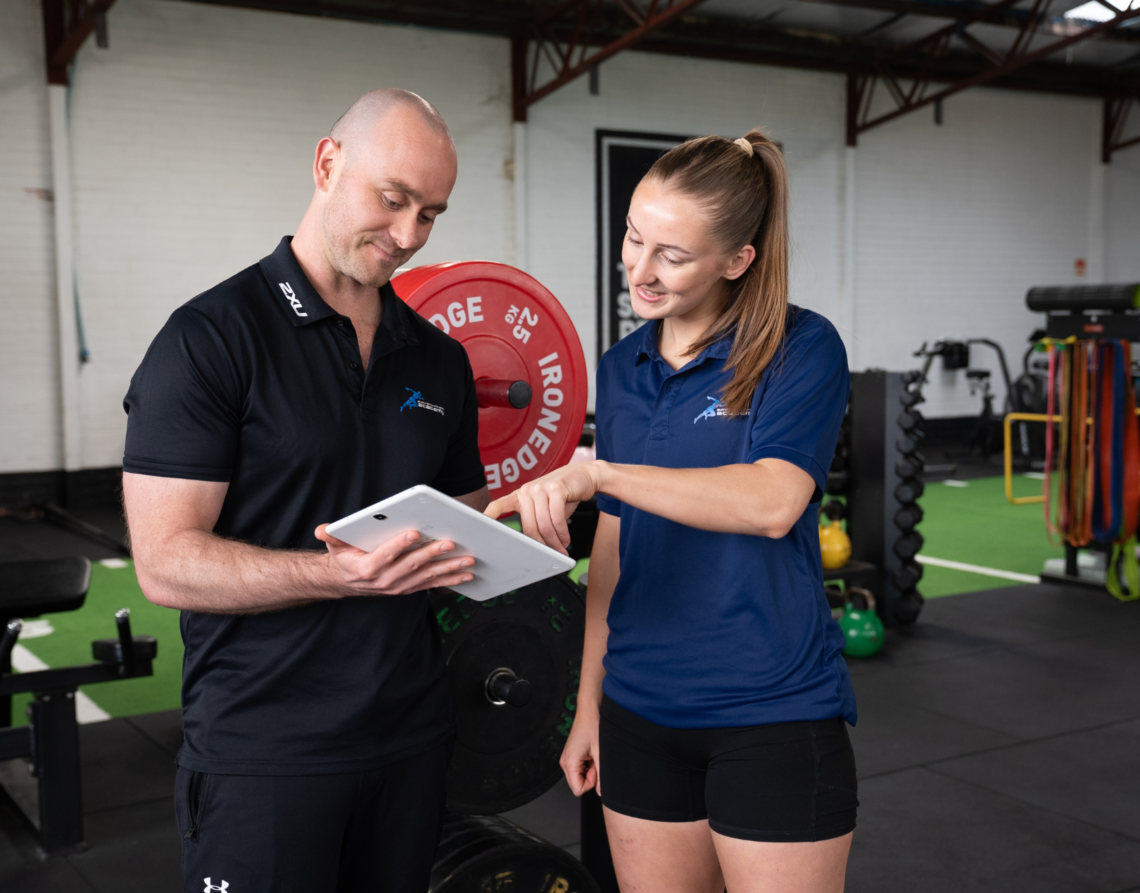 Man in black polo and woman in blue polo review a tablet together in a gym near a red Ironedge weight plate.