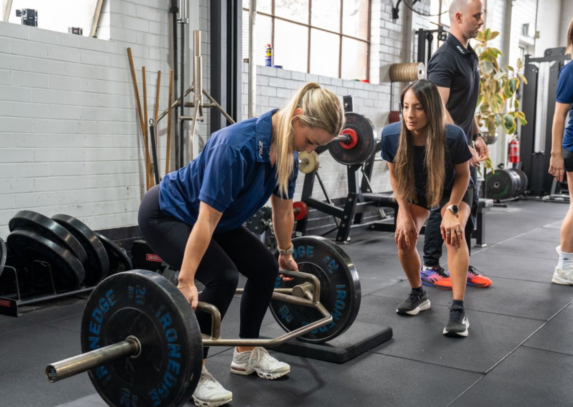 Woman in blue polo lifts a hex bar with Ironedge plates while an instructor and another woman observe in a gym.