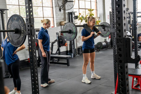 Woman in blue shirt grips a barbell on a rack while a trainer observes her form in a gym.